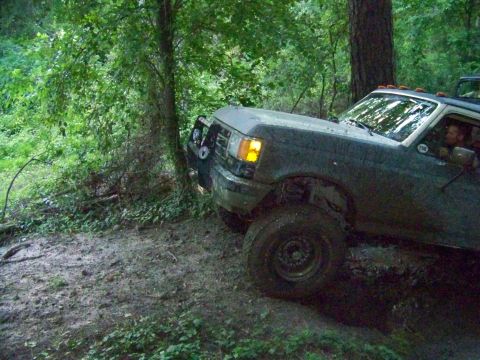 1985 Ford Bronco - A Ford F-Series truck navigates a muddy, wooded terrain.