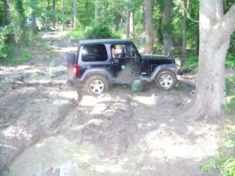 1985 Ford Bronco - A Jeep navigates a challenging muddy off-road course with trees lining the path.