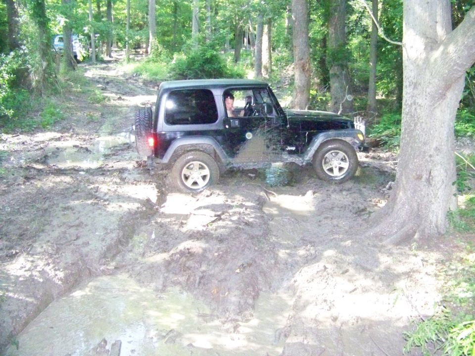 Black Jeep Wrangler driving through a muddy trail in the woods. Another vehicle is visible further up the path