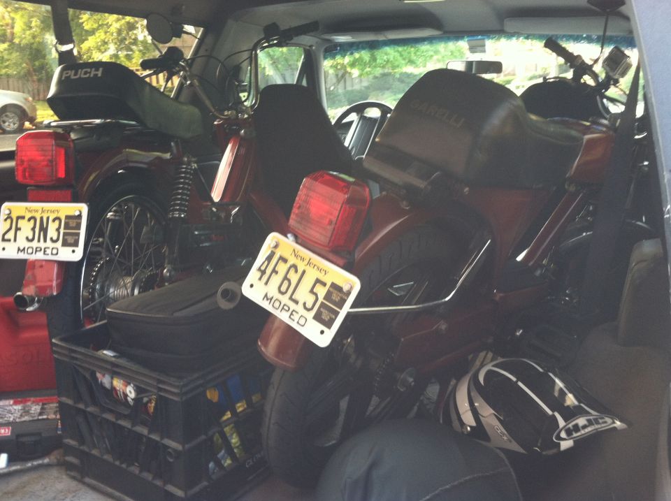 Two red mopeds loaded into the back of a vehicle. One has a PUCH logo on its cover. Both have New Jersey license plates reading 2F3N3 and 4F6L5