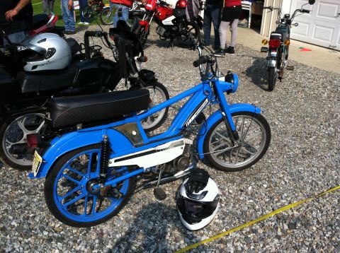 1981 Garelli Rally Sport - A blue moped and a silver helmet sit on a gravel surface, with other mopeds and people in the background.