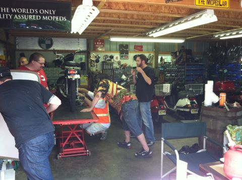 1981 Garelli Rally Sport - Several people are gathered around a motorcycle on a lift inside a cluttered garage.