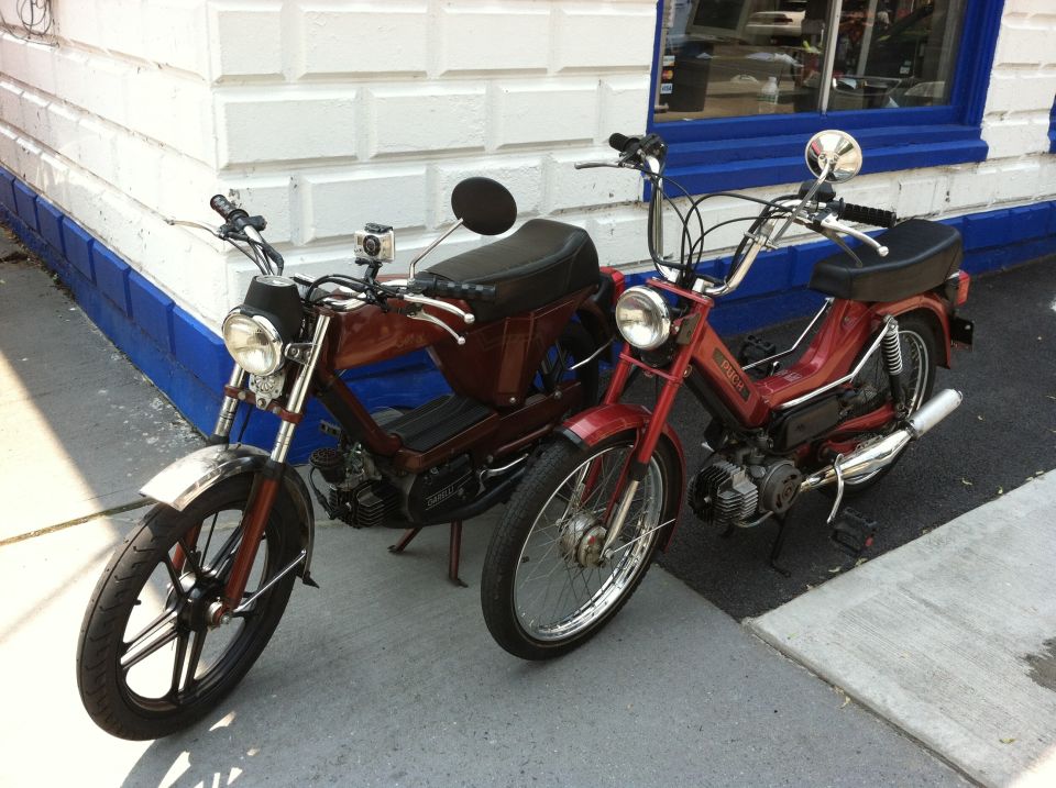 Two mopeds, one dark red and one maroon, parked on a sidewalk