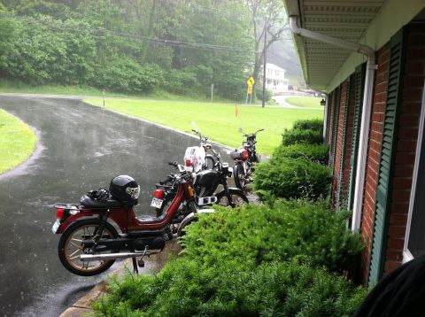 1981 Garelli Rally Sport - Mopeds are parked next to a house during a rain shower, with a wet driveway and green lawn.