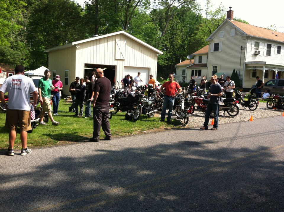 Group of people gathered around various motorcycles parked on a grassy area next to a garage and houses