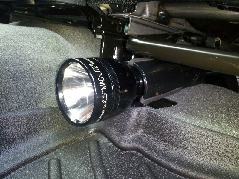 Black Maglite flashlight attached to black seat bracket, with gray carpet and black textured floor visible