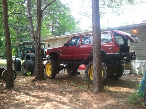 1978 Ford Bronco - A heavily modified red Jeep Cherokee sits beside a green tractor in a wooded area.