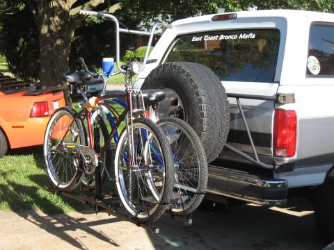 1994 Ford Bronco - White Ford Bronco with a spare tire and bicycles on a rack, parked next to an orange car.