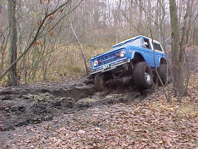 1989 Ford Bronco - A blue early Ford Bronco with a white hardtop crawls off a muddy ledge on a leaf-covered trail in bare winter woods, front driver-side whee…