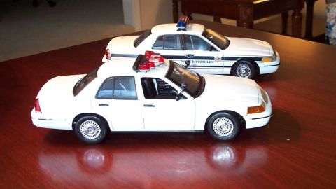 1998 Ford Crown Victoria - Two white 1998 Ford Crown Victoria police car models are displayed on a polished wooden table.