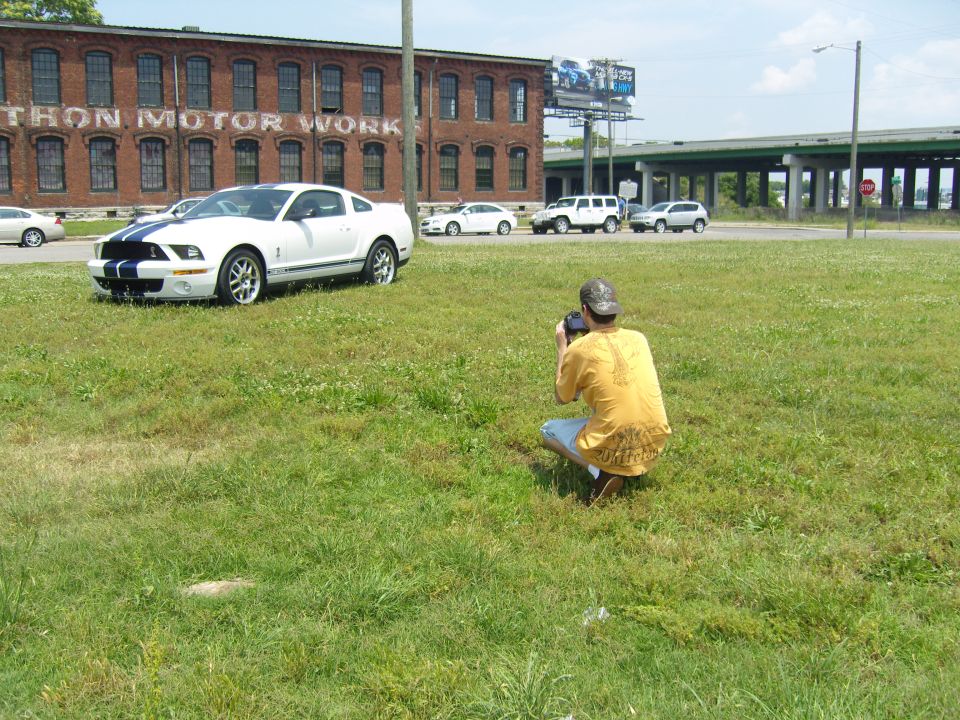 White 2007 Ford Mustang with blue racing stripes parked on grass, photographer crouching nearby
