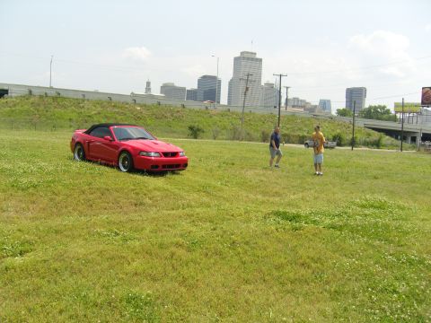 2007 Ford Mustang - A red convertible Mustang sits in a field with two men and a city skyline visible.