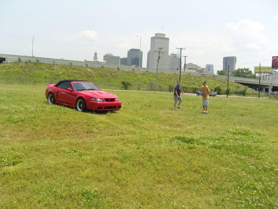 Red convertible Mustang parked on a grassy field with two men standing nearby and a city skyline in the background