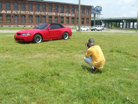 2007 Ford Mustang - A red convertible Mustang is photographed in a grassy field with an old brick building in the background.