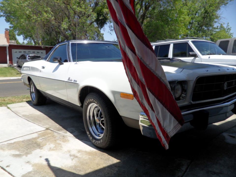 White 1973 Ford Ranchero with black hood, chrome wheels, and an American flag in front