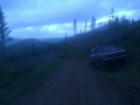 1979 Ford Bronco - A dark blue Ford Bronco sits on a dirt road with a forested hillside in the background under a cloudy sky.