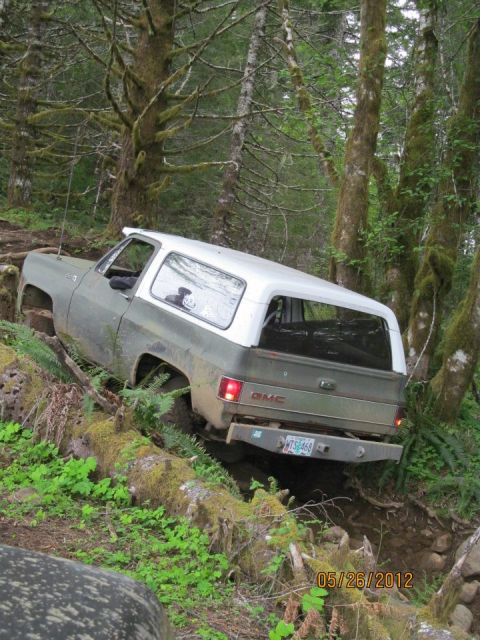 1979 Ford Bronco - A two-tone GMC K5 Blazer tackles a challenging off-road trail surrounded by dense forest.
