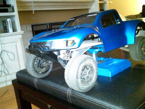 1986 Ford Bronco - A blue RC truck body is displayed on a stand, revealing its suspension and off-road tires.