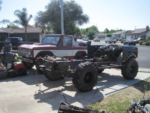 1989 Ford Bronco - A Ford Bronco body sits next to its disassembled chassis and engine, with other vehicles and houses in the background.