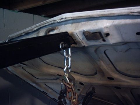 1991 Ford LTD Crown Victoria - Low-angle shot of a white body panel's ribbed underside with a black engine hoist or lift arm hooked to it by a shackle and chain link. Bar…