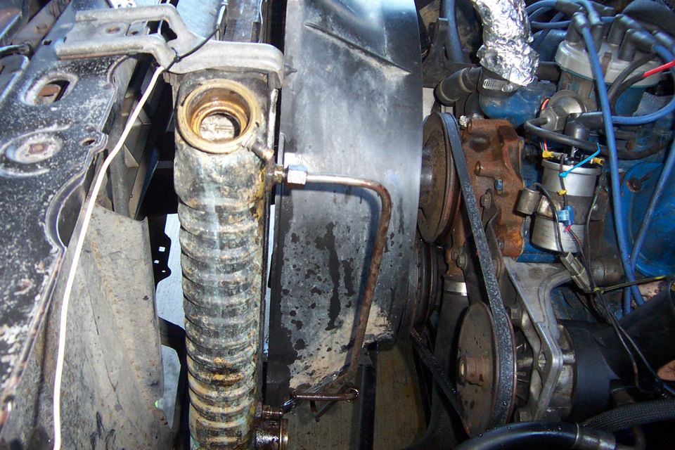 Open radiator filler neck with scaling next to transmission cooler line and engine pulleys