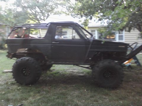 1986 Ford Bronco - Modified black Ford Bronco with oversized tires and a roll cage sits in a grassy yard.