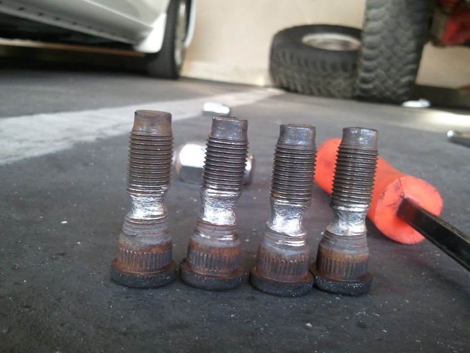four used wheel studs on a dark surface, with a tire and part of a car in the background