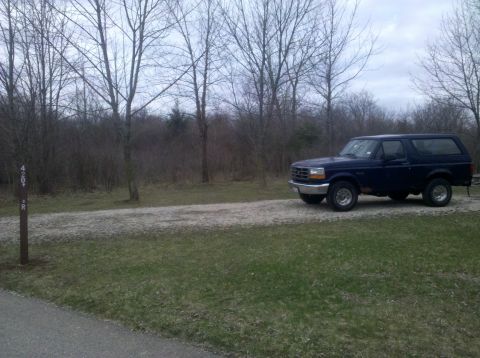 1995 Ford Bronco - A dark blue 1995 Ford Bronco is parked on a gravel road surrounded by bare trees.