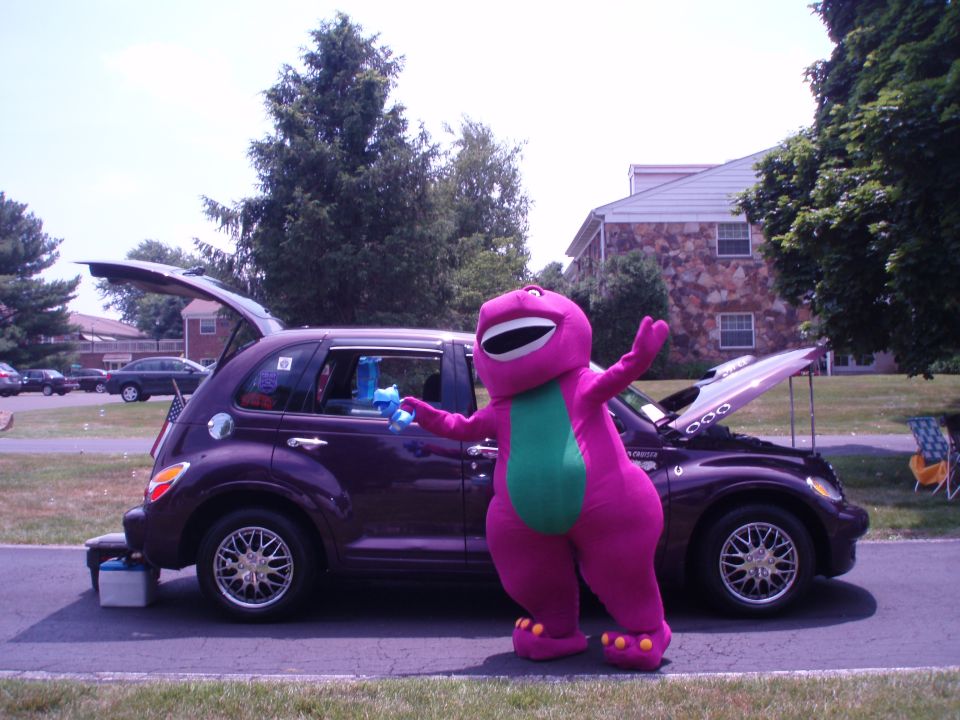 Person in purple Barney dinosaur costume washing a dark PT Cruiser with hood and hatch open