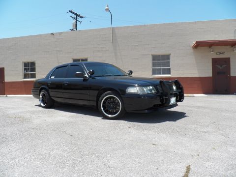 2003 Ford Crown Victoria - Front left three-quarter of a black Crown Victoria with brush guard and illuminated headlights.
