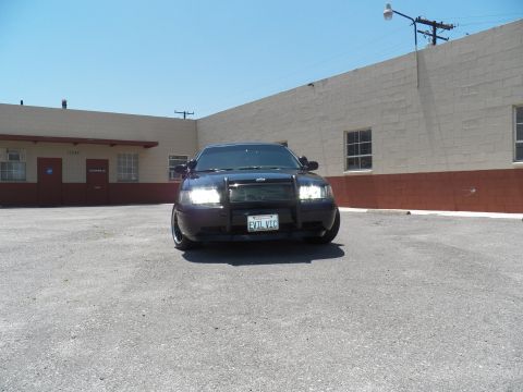 2003 Ford Crown Victoria - Front shot of a lowered black Crown Victoria sedan, headlights on, parked in a paved lot by a tan building.