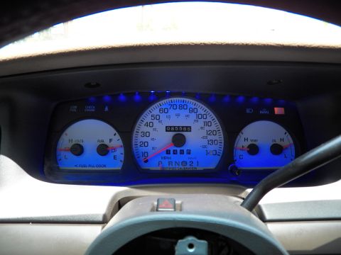 2003 Ford Crown Victoria - Over-the-wheel view of the blue-glowing instrument cluster in a tan dash, hazard button visible.