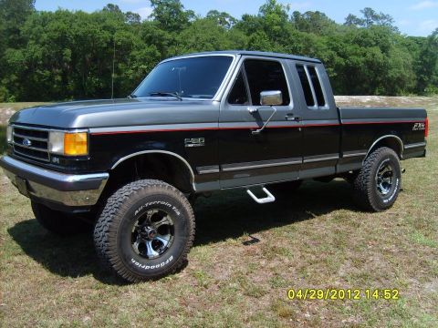 1989 Ford F-150 - This lifted Ford F-150 pickup truck features a two-tone paint scheme and off-road tires, parked outdoors on a grassy area.