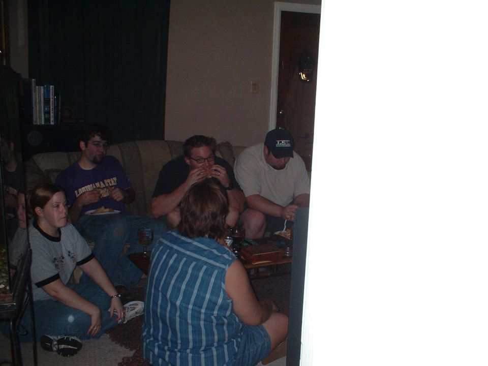Group of people eating in a dim living room, one wearing an LSU cap