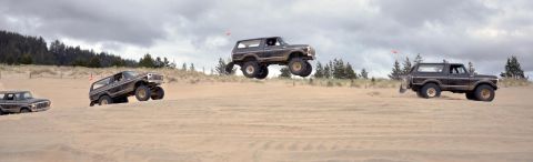 1979 Ford Bronco - Multiple Ford Broncos are seen off-roading and jumping over sand dunes on a cloudy day.