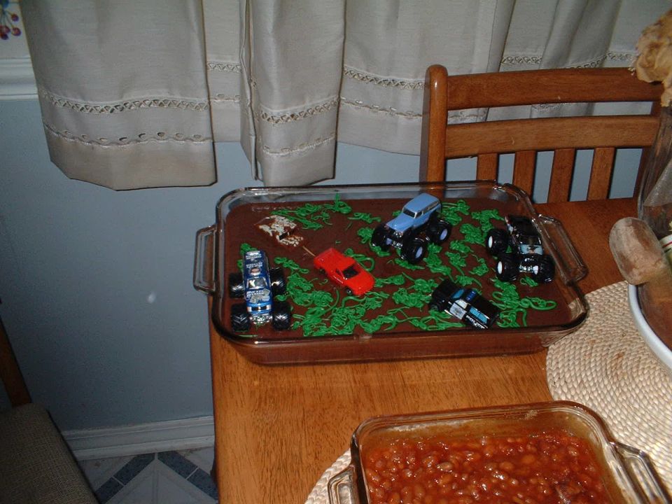 Chocolate sheet cake decorated with toy monster trucks next to a dish of baked beans