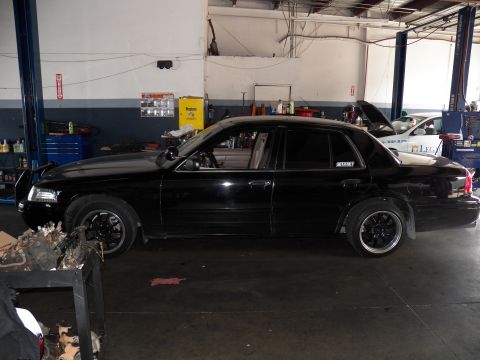 2003 Ford Crown Victoria - Driver-side profile of a black Crown Victoria inside a repair shop, lowered on dark multi-spoke wheels.