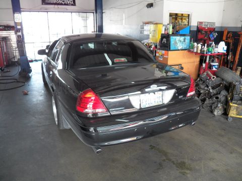 2003 Ford Crown Victoria - Rear three-quarter of a black Crown Victoria inside a busy shop with shelving and engine parts behind.