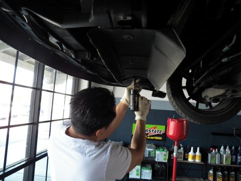 2003 Ford Crown Victoria - Worker in gloves operates a tool on the underbody of a raised vehicle beside a red oil funnel.