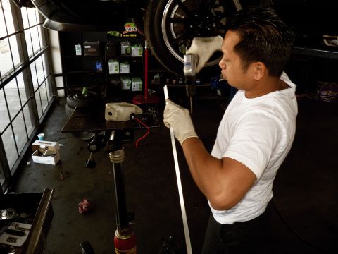 2003 Ford Crown Victoria - Worker in gloves drills into a driveshaft held on a stand, wheel and tools above and around him.