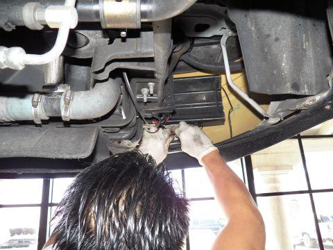 2003 Ford Crown Victoria - Mechanic reaches up to wire a component beside coolant hoses and an exhaust under a raised car