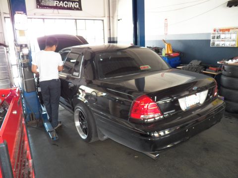 2003 Ford Crown Victoria - Rear quarter of a black Crown Victoria, hood raised, mechanic working over the engine