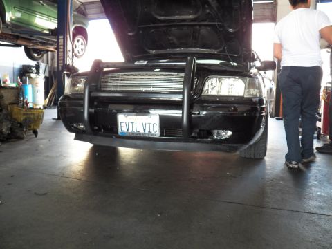 2003 Ford Crown Victoria - Black Crown Victoria front with push bar and EVIL VIC plate, worker standing at right