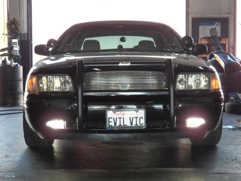 2003 Ford Crown Victoria - Black Ford Crown Victoria, front view with push bar, fog lights lit, California plate reading EVIL VIC, in a shop.