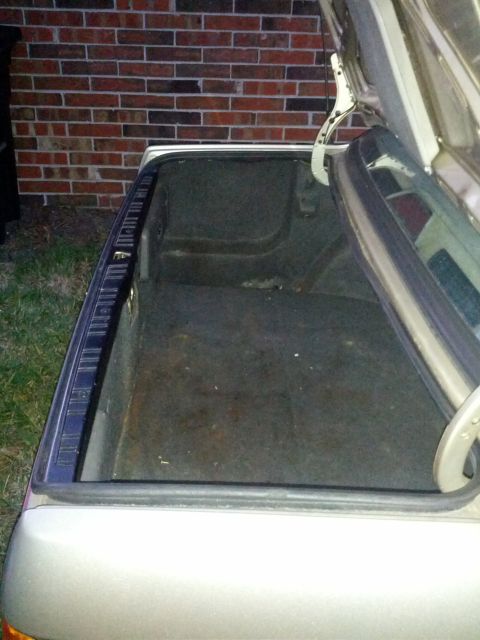 1989 BMW 3 Series - The interior of an open car trunk is visible, with a brick wall and grass in the background.