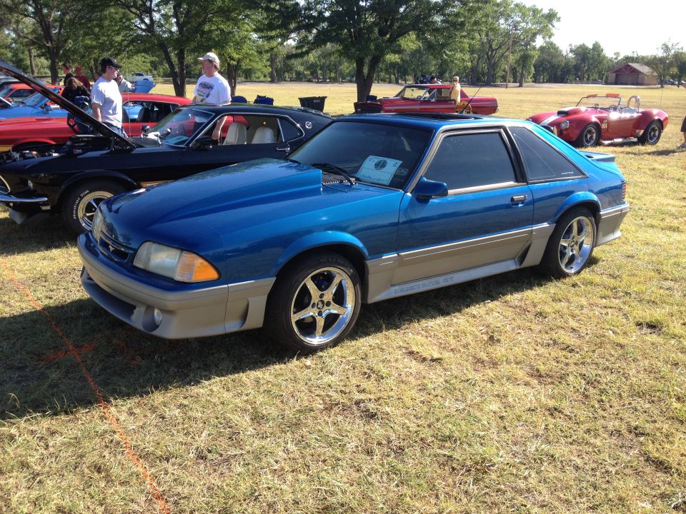 Blue 1990 Ford Mustang with a grey lower body kit and aftermarket wheels, parked on grass