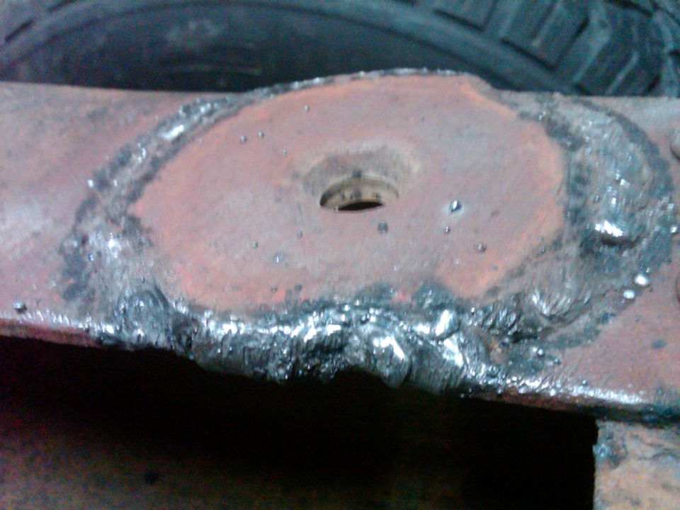 Close-up of a rusty metal part with rough welding around a central hole. A tire is visible in the background