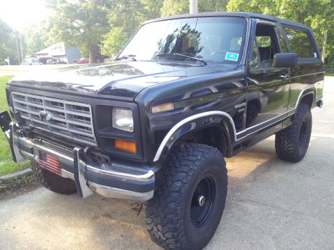 1986 Ford Bronco - This black Ford Bronco is parked on a driveway with trees and houses in the background.
