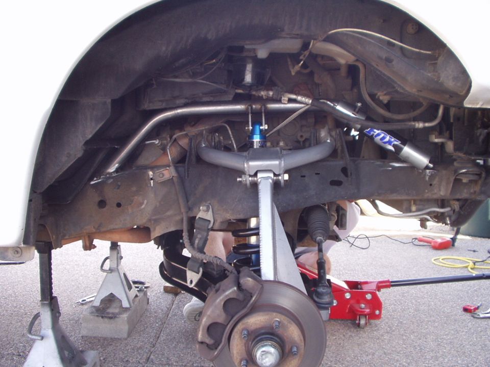 Front suspension underside with coil spring, brake rotor, and blue FOX shock on a lifted white truck