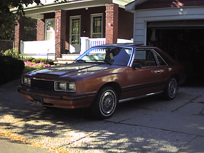 Brown Mercury Capri hatchback parked in driveway in front of brick house with porch